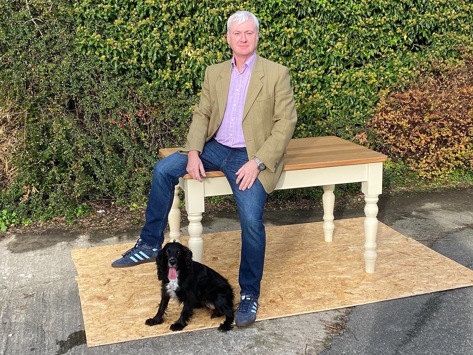 Neil sitting on a finished table he made, with a painted base, turned legs and a solid oak top, and a spaniel at his feet